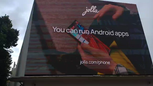 Eine große Werbeleinwand an der Wand in Organge gehalten. Oben das Jolla Logo darunter eine Szene wo ein Finger den Touchsceen des Jollaphone berührt. Dazu eracheint der Text You can run Android Apps... In der nächsten Einstellung ein Finger der einen Hardwareschalter an der Seite berührt. Dazu die Texteinblendung "Or shut them off if you don't trust them" In der nächsten Einstellung das Telefon aus drei Seiten mit der Texteinblendung "My phone. My Data."