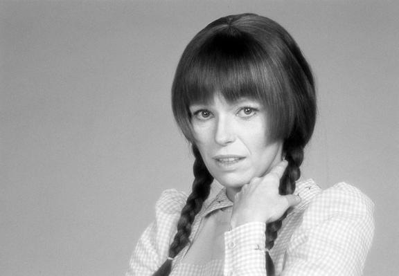 Black and white portrait of a woman with bangs and two braided pigtails, wearing a collared checkered shirt, with her hand resting on her neck.