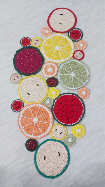 Crochet table runner laid out on a white surface. It's made of several different circles varying in size. Each circle represents a piece of fruit, there are apples, strawberries, watermelon, lemons, oranges and limes.