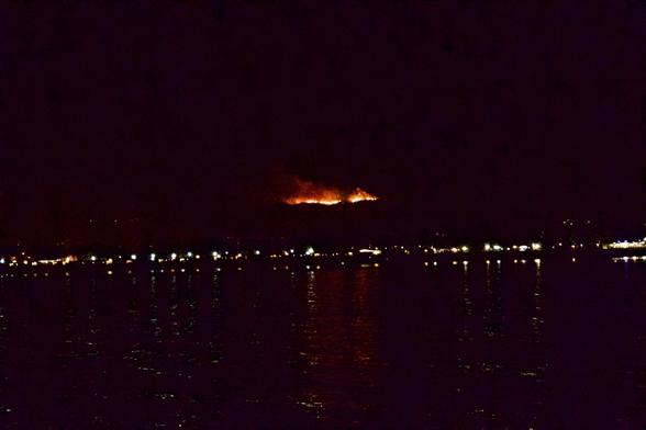 Forest fire burning its way down a mountain in a line of fire towards the lights on shore at night.