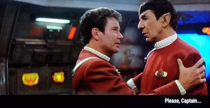Star Trek The Final Frontier scene. Kirk and Spock, friendly colleagues, are on the bridge in their 'movie red' uniforms. Kirk is facing Spock and holding onto his shoulders as if he's going to share a long kept secret that dareth not be spoketh.... or maybe just going in for a hug, as good work associates do. Spock is just standing and speaking, and being held by Kirk. Closed caption reads, "Please, Captain... Not in front of the Klingons."