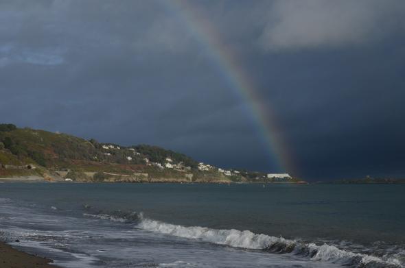Ein Landschaftsfoto einer Küste unter einem dunklen Himmel mit einem Regenbogen. Im Vordergrund brechen kleine weiße Wellen an einem dunklen Sandstrand. Das Meer erstreckt sich bis zu einer hügeligen Küstenlinie auf der linken Seite, die mit grüner Vegetation und vereinzelten weißen Gebäude Gebäuden bewachsen ist. Ein Regenbogen zieht sich diagonal von oben in der Bildmitte durch den Himmel nach rechts unten. Der Himmel ist überwiegend dunkelblau und grau mit einigen helleren Wolken im oberen Bereich.