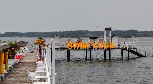 Ein Steg mit Tischen und Stühlen führt zu einem "Swim Club" auf einem Steg mit Strandkörben und Dekorationen.