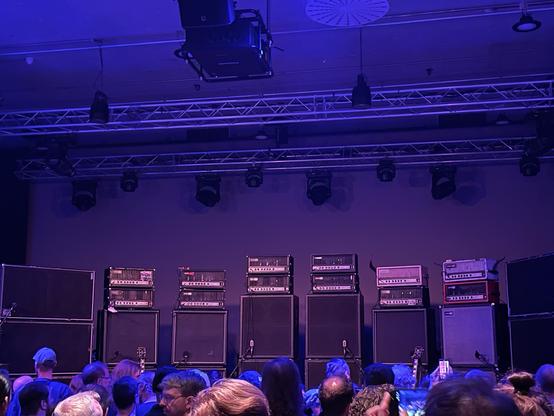 Auf einer Bühne stehen 8 Türme mit Gitarrenverstärkern und Lautsprechern. Über der Bühne befinden sich Traversen mit ein paar Scheinwerfern, vor der Bühne ist Publikum zu erkennen.