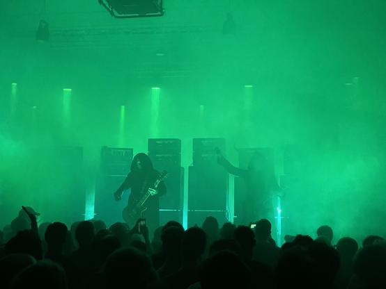 Zwei Gitarristen die in Mönchskutten gekleidet sind stehen in grünem Nebel vor den 8 Verstärkertürmen.