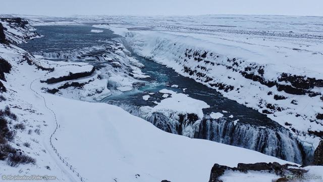 Four colour photos of parts of a frozen waterfall under a snow-laden sky. In the upper left of this shot a wide blue river is making its way towards the viewpoint. Near the centre right it drops over a clutter of eroded boulders and steps, many of which are covered in snow and ice. The canyon wall next to this upper falls is caked with ice. The water course then turns ninety degrees to another drop into a deep canyon, just in front of the viewer. The foreground is a snow field with a large cornice overhanging the canyon, although this isn't obvious from this angle. A line of fencing is running down to a natural rock platform next to the top falls. Compacted ice shows where people have walked down.