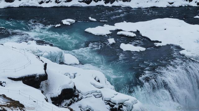 A close-up of the crest of the drop into the canyon. The swirling blue water is white with foam as it passes the natural rock platform which is closed to visitors. Zooming in on the right to 100% of the photo shows footprints all around the edge where visitors have ignored the safety warnings. Icicles and clumps of heavy turquoise ice have formed along the edge of the platform and the snow shows distinct layers.