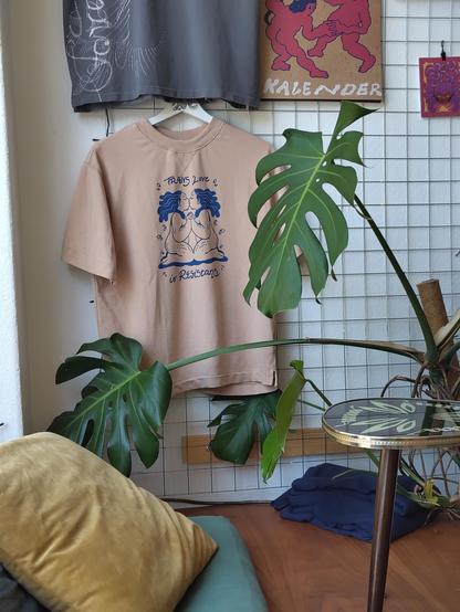 a t-shirt hanging in a cafe behind a monsterra, it is two Black trans people holding hands on their knees and kissing. it says trans love is resistance