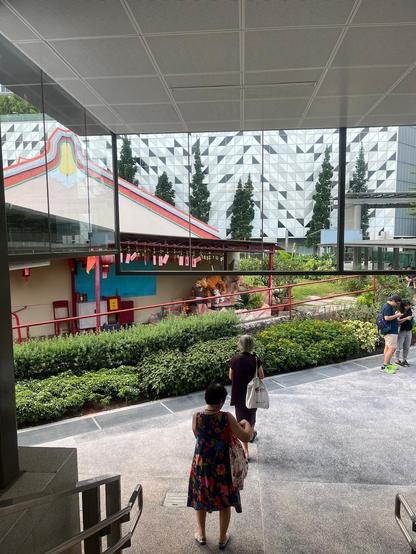 People walking through a modern outdoor courtyard with a temple-like building, greenery, and a tiger statue.