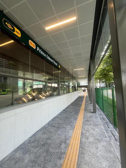 Covered walkway at Prince Edward Road MRT station, with glass walls, tactile paving, and trees outside.