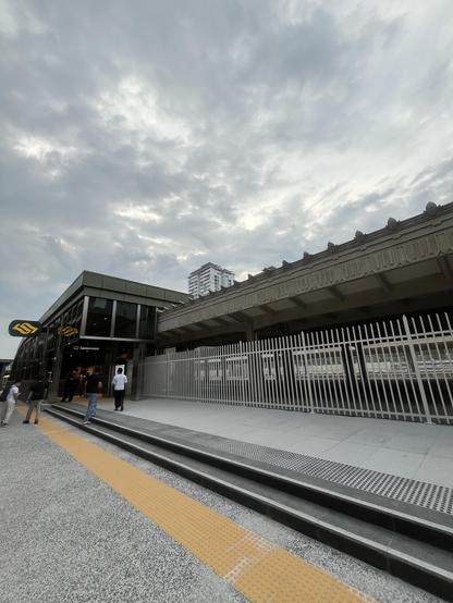 Exterior of a modern train station with a cloudy sky, people walking in, and a fenced platform area.