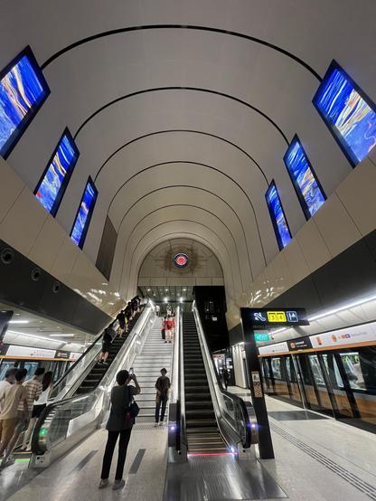 A modern subway station interior with a high arched ceiling, blue digital wall displays, and people riding escalators.