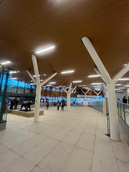 Modern covered walkway with white tree-like columns, ceiling lights, a few people, and bronze animal sculptures.
