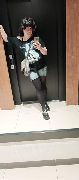 Mirror selfie of me, a blackhaired transfem of 39, looking gorgeous and striking a pose that shows Attitude, leaning against a wall and showing off my legs. I'm wearing short blue jean shorts over opaqueish black tights, a Nightwish T-shirt cut up to show off one shoulder over a black bra, and black men's Mary Jane shoes adorned with chains. I have a gray fuzzy bag slung over my shoulder. I look basically like a rock star with my hair that I've carefully cut to look wild and unruly.