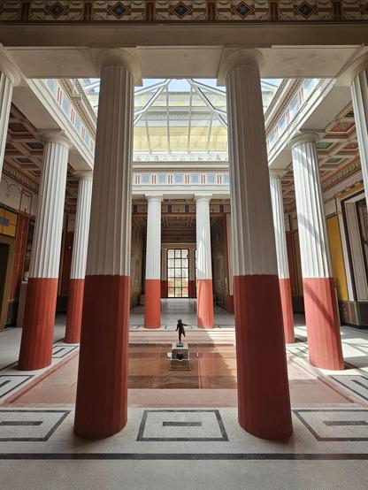 A bright Roman style atrium (open central court with tall white-and-red columns, a skylit ceiling, and a sgallow basin with a small bronze statue at the center.
