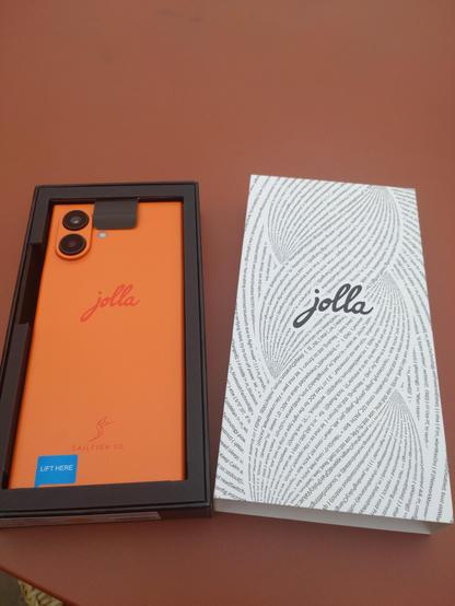 Ein Jollaphone in der Verpackungsbox liegend. Das Telefon hat ein Oranges Backcover mit Jolla Schriftzug.