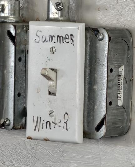 A light switch is labeled in marker "Summer" and "Winter". The switch is currently set to "Summer". 

It is a western-style toggle light switch on a metal surface junction box with metal conduit coming out the top. A second outlet box is to the left, but the image is cropped so that we can't tell what is in that box.The white switch and faceplate are both somewhat dirty. There's some suggestion of an invisible pink unicorn off to the side.