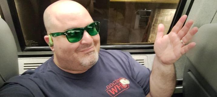 Photo of the Undersigned sitting on a train seat, wearing green sunglasses and a Doctor Who t-shirt, smiling and gesturing toward the window behind him through which the inside of a train station is visible.