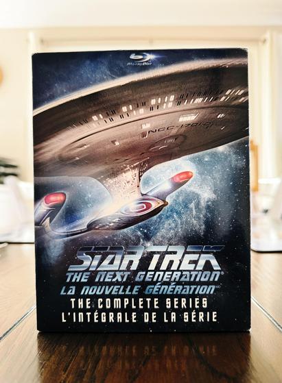 A photo of the box of the Star Trek: The Next Generation "The Complete Series" Blu-ray box set. On the front it has a dramatic photo of the U.S.S. Enterprise D with a blue nebula/starry background behind it, and underneath the English text is French translation.