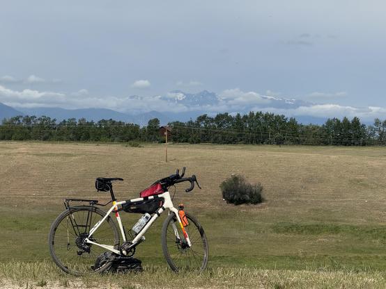 A white gravel bike parked on the edge of a hill. A line of cottonwood trees along an irrigation ditch crosses the far side of The pasture behind the bike. Low storm clouds break against the northern slopes of the Beartooth Mountains in the background. The early July storms and cool weather are a pleasant surprise.
