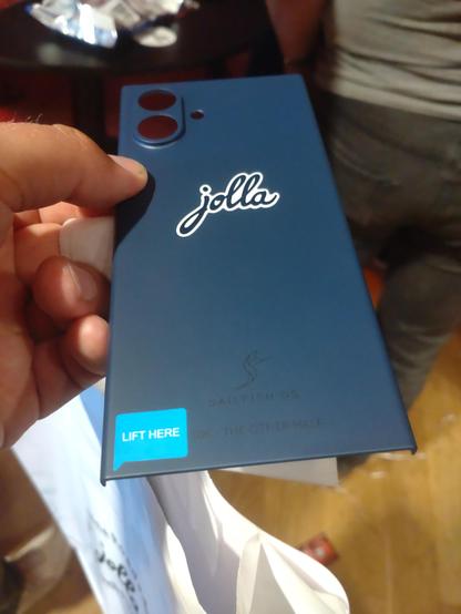 Eine Rückseitenschale für das Jolla Phone in einem tiefen Blauton. Ein großes weißes Jolla Logo ist darauf zu sehen. In der linken Ecke unten ist ein Aufkleber "Lift here" angebracht. In weniger auffälliger Schwarzer Farbe ist noch eon SailfishOS Logo und der Schriftzug 10K THE OTHER HALF angebracht.