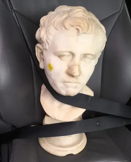 A Roman marble portrait of a man strapped into a black car seat with two seatbelts crossing over it.