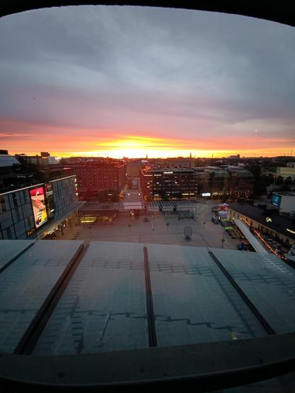 Ein Sonnenuntergang über Helsinki. Der Blick fällt auf den Platz vpr dem Kamppi. An dem Gebäude vom Kamppi ist eine große LED Werbewand angebracht auf der gerade Werbung für das Jolla Phone zu sehen ist.