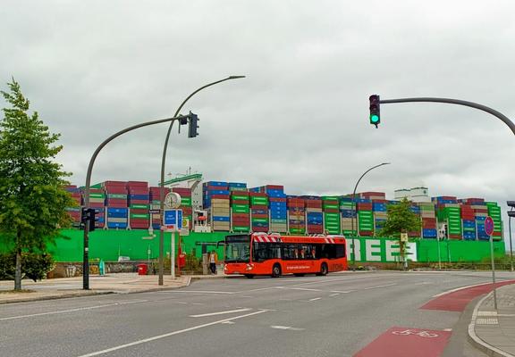 Auf der anderen Seite einer Straße steht ein Bus in orangerot und Baustellen-Optik mit rot-weißen Streifen am Dach. Hinter ihm fährt ein grünes Containerschiff mit vielen geladenen Containern.