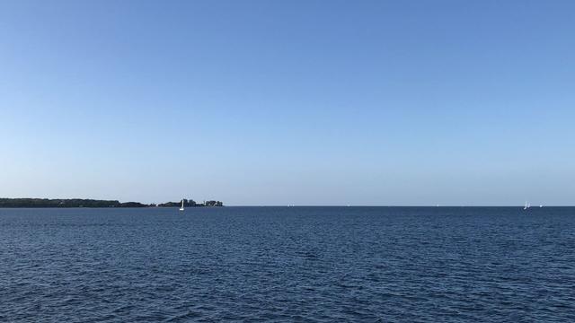 Dunkelblaues Wasser, hellblauer Himmel, heller am Horizont. Links eine Landspitze mit Leuchtturm zwischen Bäumen.