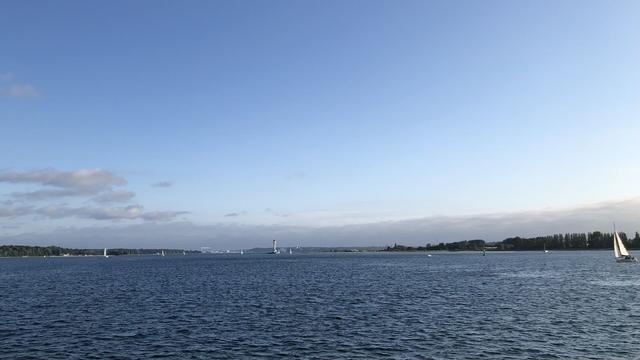 Andere Richtung, weit entfernt im Wolken-Dunst die Stadt. Rechts am Rand ein Segelboot im Sonnenlicht.
