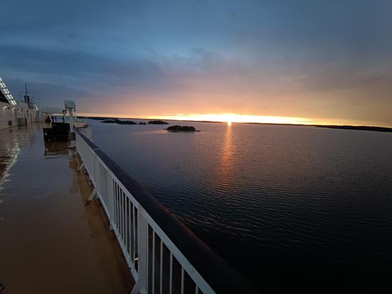 Foto aufgenommen mit der Weitwinkellinse. Blick in den Abendhimmel auf See in den Finnischen Schären. Die Sonne bricht gerade unter die Wolkendecke, die Wolken am Himmel in dunklem Stahlblau setzen einen tiefen Kontrast zum Orange der Sonne. Im linken Teil des bildes ist das Sonnendeck der Fähre zu sehen. Das Deck ist sehr naß es hat geregnet.
