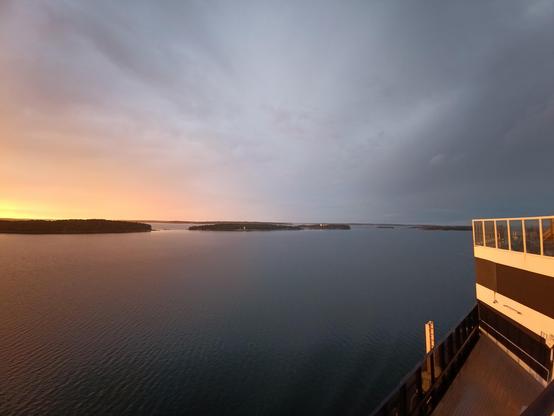 Blick Achtern ab. Der  Bereich wo die Sonne untergeht ist nur partiell am linken Rand im Foto aufgenommen und erscheint mit vielen Orange und Rottönen am linken Horizont. Darunter sind einige bewaldete Schären zu sehen. Der Himmel hat einen tiefen Kontrast in Stahlblauen Tönen. Das Wasser liegt leicht gekräuselt. Am rechten Rand sieht man in warmen Sonnenlicht beschienen einen Teil der Fähre. Die Aufnahme ist ebenfalls mit der Weitwinkel Kamera erfolgt.