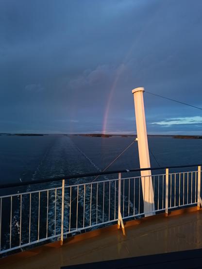 Aufnahme vom Heck des Schiffes ab. Auf dem Wasser lässt sich durch leichte Schaumkronen erkennen wo das Schiff gerade entlang gefahren ist. Am Himmel steht ein Regenbogen mit seinem Ende auf einer Schäreninsel. Im Vordergrund ist die Reeling der Fähre zu erkennen die von warmen Sonnenuntergangslicht beschienen wird.