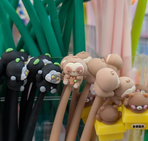 SLOTH PEN

A light brown silicone-encased pen topped by an artistic chibi interpretation of a sloth gripping a small tree branch, in a store display surrounded by many other pens of identical and similar designs