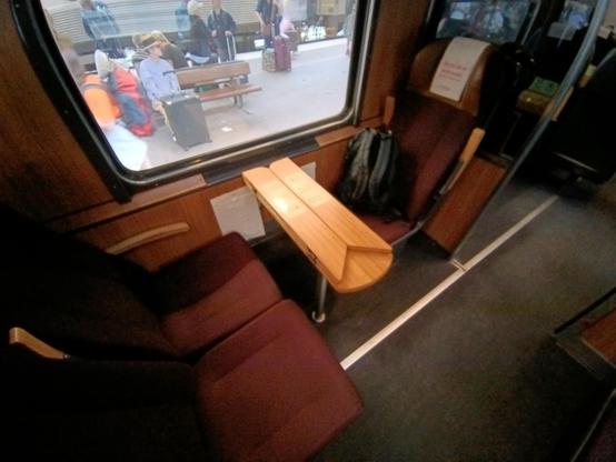 Eine 3er vis 'a vis Sitzgruppe mit Tisch. Der Rucksack ist auf den Einzelplatz abgelegt. Die Einrichtung sieht gemütlich aus. Die Sitze sind mit einem Weinroten Bezug ausgestattet. Die Wände mit einer Holzfunieroptik. Der Tisch ist wie sonst oft in der 1. Klasse üblich durch ausklappen zu vergrößern.
