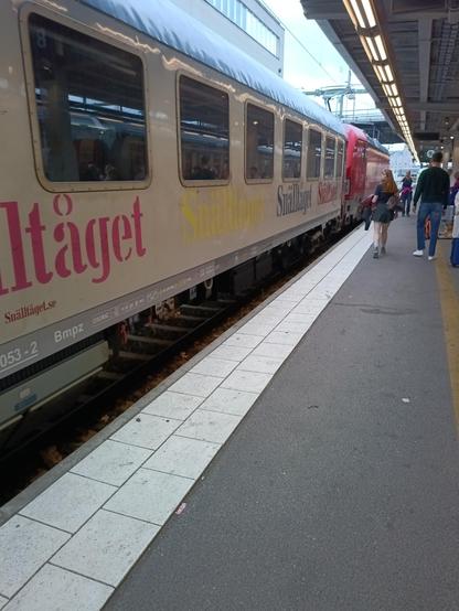 Bild vom Bahnsteig wo der mit bunten Schriftzügen versehene Snälltåget einfährt. Er wird von einer roten Lok gezogen