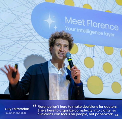 White male with curly hair (CEO of LongevityAI, HQ in Israel, Guy Leitersdorf) holding a microphone near mouth, standing before a backdrop of interconnected dots and white text in a blue bubble that reads: Meet Florence your intelligence layer.