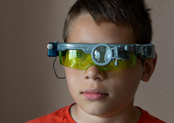 Close-up portrait of a young boy wearing futuristic yellow-tinted toy goggles with a magnifying lens and a small blue light. (AltTexty)