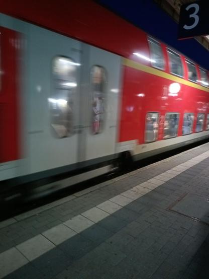 Eine Regio Roter Doppelstockzug fährt am Bahnsteig ein. Der Zug hat eine Bewegungsunschärfe auf der Aufnahme. Davor ist der Bahnsteig zu erkennen und eine 3 die das Gleis 3 kennzeichnet die von der Decke gehangen ist.