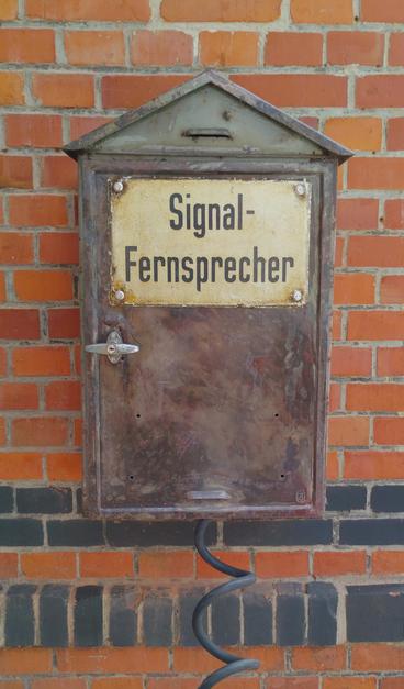 Zu sehen ist ein rostiger Metallkasten vor einer einer roten Backsteinmauer, aus dessen Unterseite ein Kabel Richtung Betrachter abgeht und auf dessen Vorderseite ein goldgelb glänzendes Metallschild mit dem schwarzen Schriftzug Signal-Fernsprecher prangt.

Der rostige Kasten hat ein spitzes Dach und die Mauer in Hintergrund enthält unten im Bild zwei horizontale, schwarze Backsteinzierreihen.