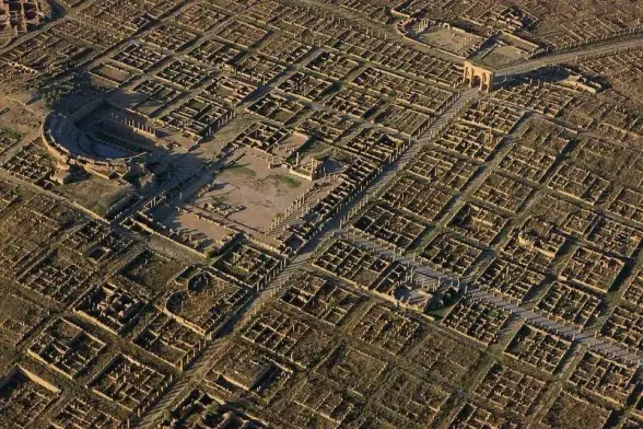 An aerial view of the extensive ruins of a Roman town, showing a well-preserved grid layout with stone foundations, streets, and colonnades. Visible landmarks include a large amphitheater, a central forum surrounded by columns, and monumental arches, all set within a geometric network of buildings and thoroughfares.