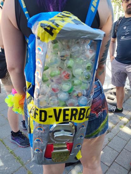 Ein Kaugummiautomat mit Münzeinwurf und Drehknauf, der mithilfe einer Ikea-Tüte und afd-verbots-Flatterband zu einem Rucksack umgebaut wurde.