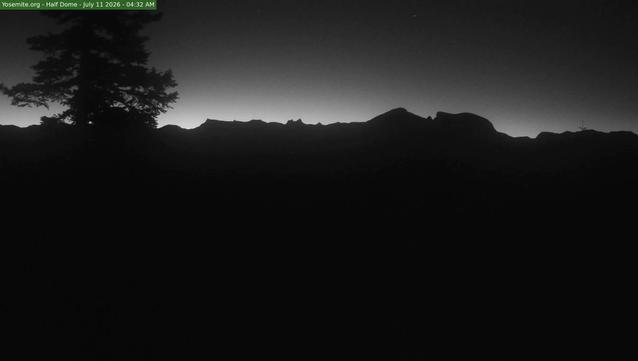 A landscape view of the peaks and domes of Yosemite Valley at sunrise, with the sky a dim gradient from black to bright white, starkly contrasted on just the outline of unicorn peak, the cockscomb, tressider peak, cathedral peak, clouds’ rest, and half dome. A large pine tree is silhouetted in the left third of the frame.