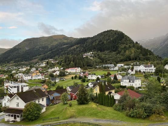 A picturesque small Norwegian village with a smallish mountain in the background. White and grey clouds in the sky to the right with clearing skies to the left. Dozens of scattered houses populate the foreground.