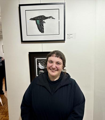 A non-binary person with a goofy grin and a sick as haircut of microbangs and goth tendrils standing in a small gallery in front of a linoprint of a Pacific Black Duck.