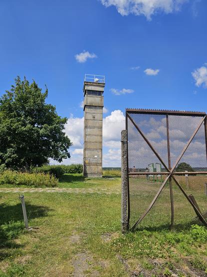 A tall concrete watchtower stands in a grassy field with a large tree to the left and a metal fence in the foreground on the right, beneath a bright blue sky with scattered clouds.