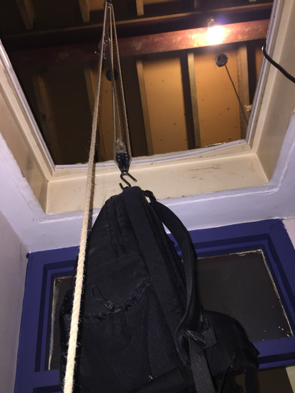 A typical loft hatch - a hole in the ceiling of a hallway leading up to the roof void. Through the hatch dangles a gun tackle - a lifting arrangement consisting of two pulleys and a rope linking them, with a hook hanging from the bottom pulley; a backpack is suspended from the hook.