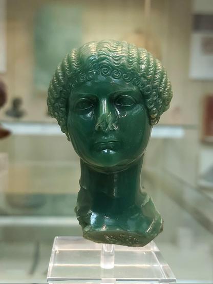 A green stone bust of a woman displayed in a museum. The sculpture features an elaborate hairdo and a solemn expression. The bust is mounted on a clear stand, with a blurred background showcasing other artifacts in the exhibition.