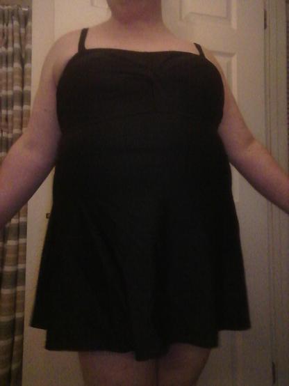 A pale woman in a black swimsuit facing the camera. Her arms are spread.