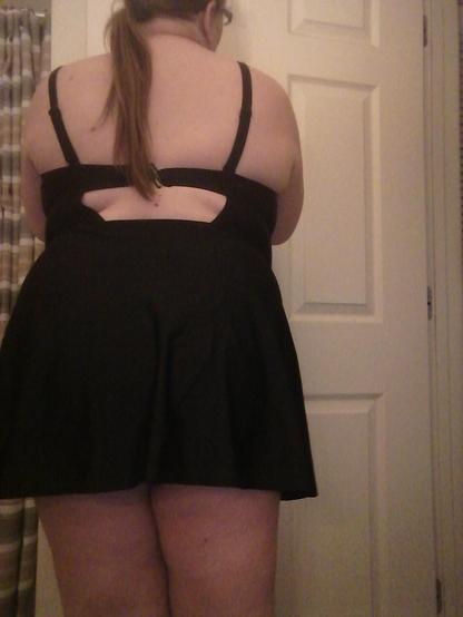 A pale woman in a black one-piece swimsuit facing array from the camera. The bra band portion of the suit is squeezing the soft fat of her sides and back, forming a muffin-top over it.